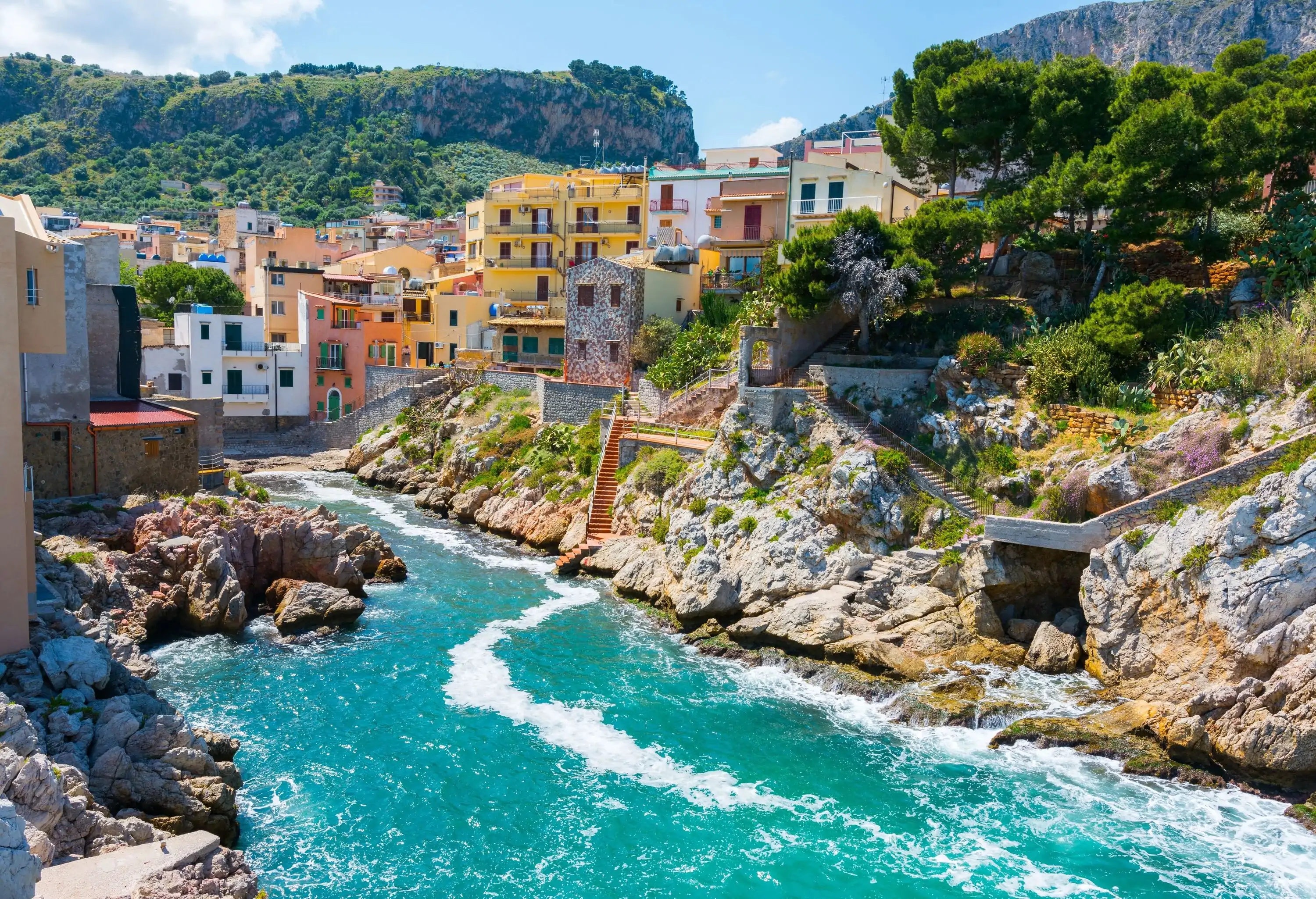 A narrow bay with colourful houses and trees along the rough shoreline.