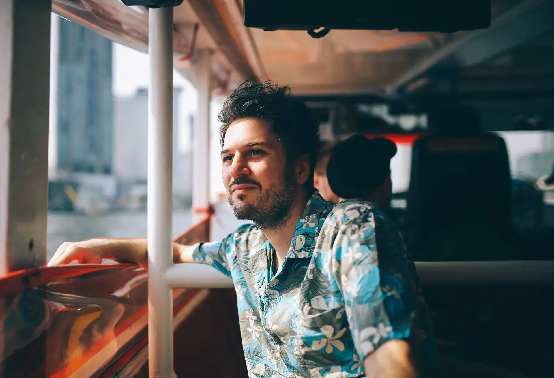 Young tourist moving around Bangkok, using a ferry boat. He is wearing a summertime fashion Hawaiian shirt, watching the Bangkok panorama around him.
