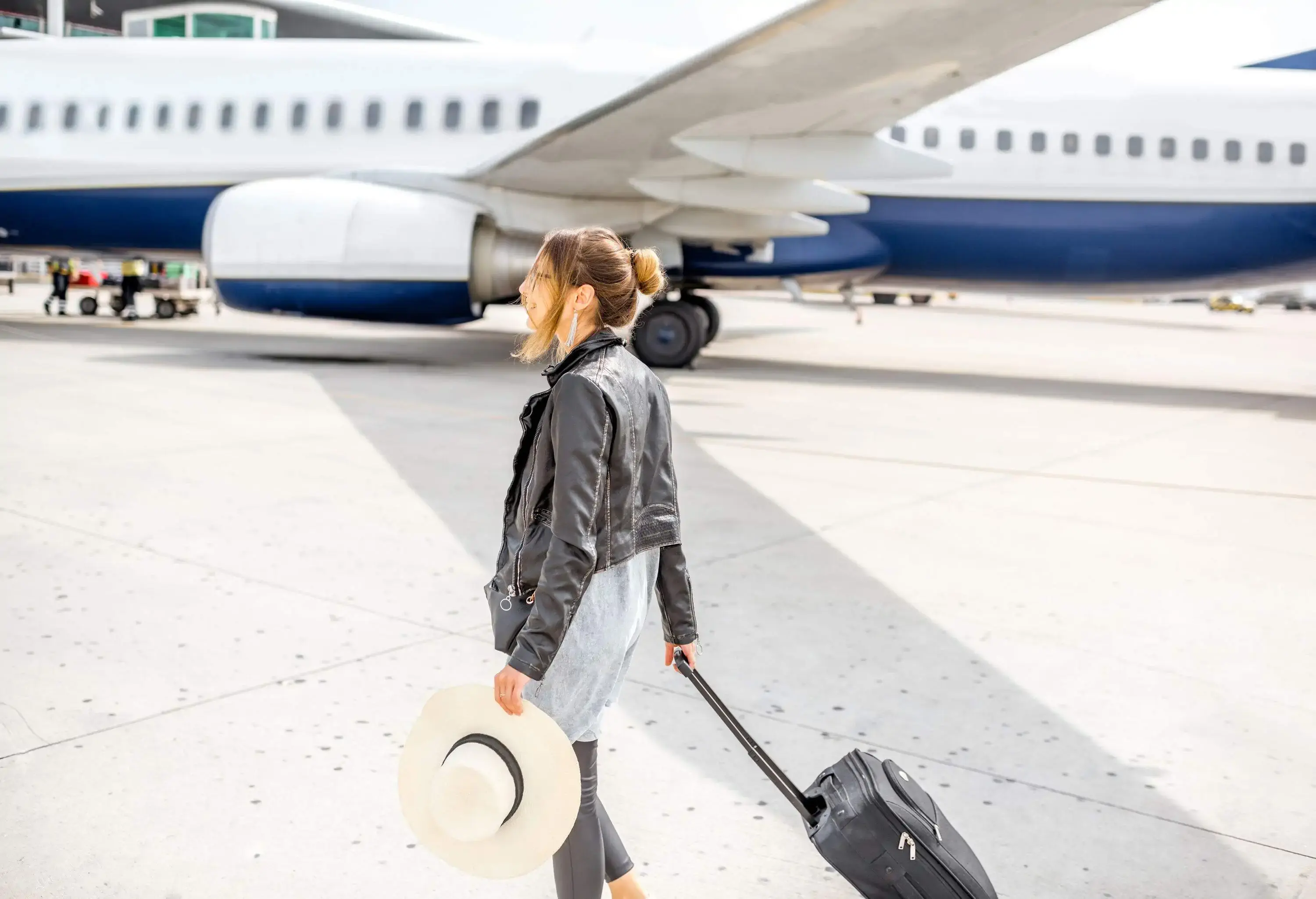 A woman in a black jacket hauling her bag across the tarmac while holding a large hat. 