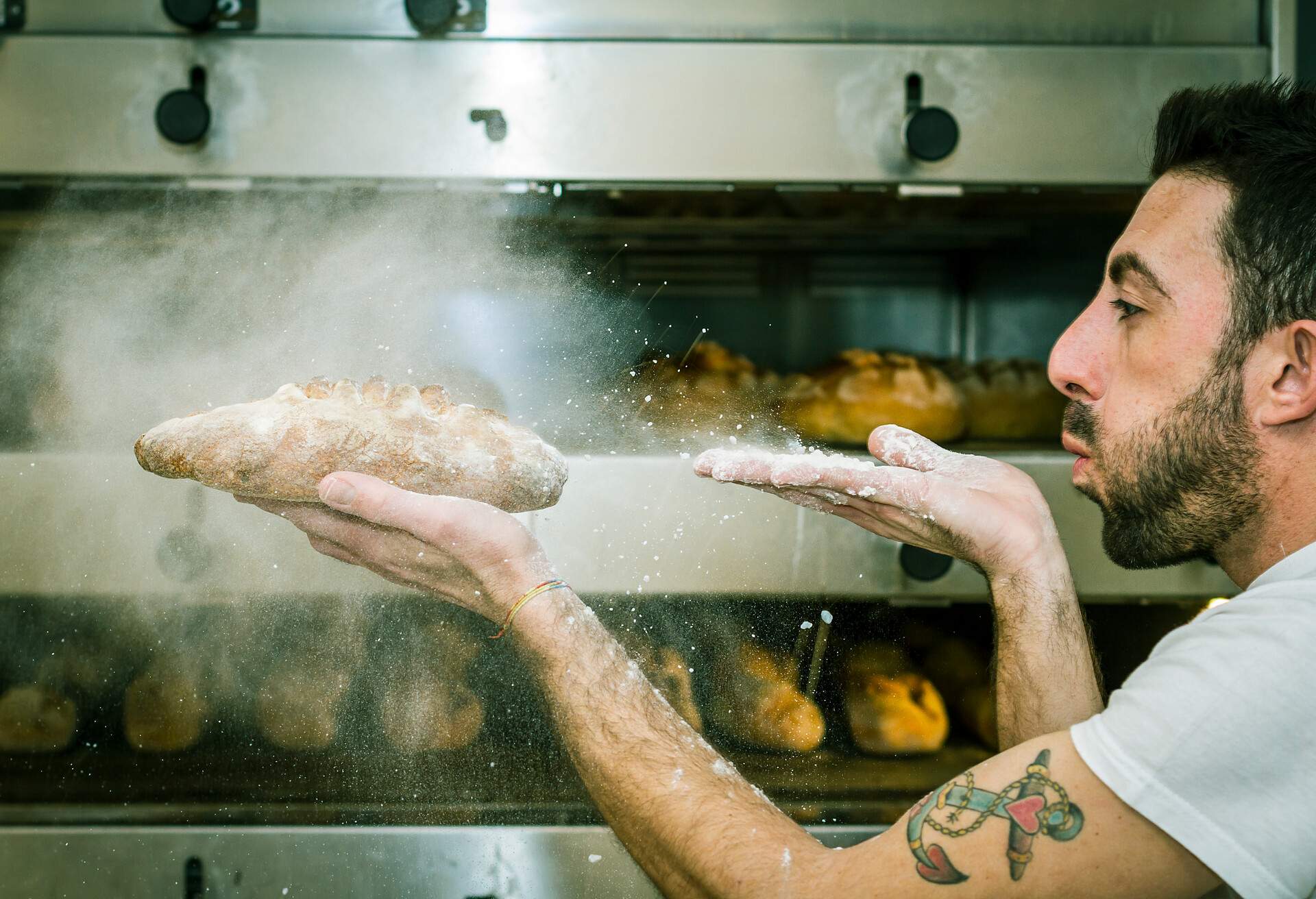 Young man (baker) blows the flour on bread - Oven with many breads on background