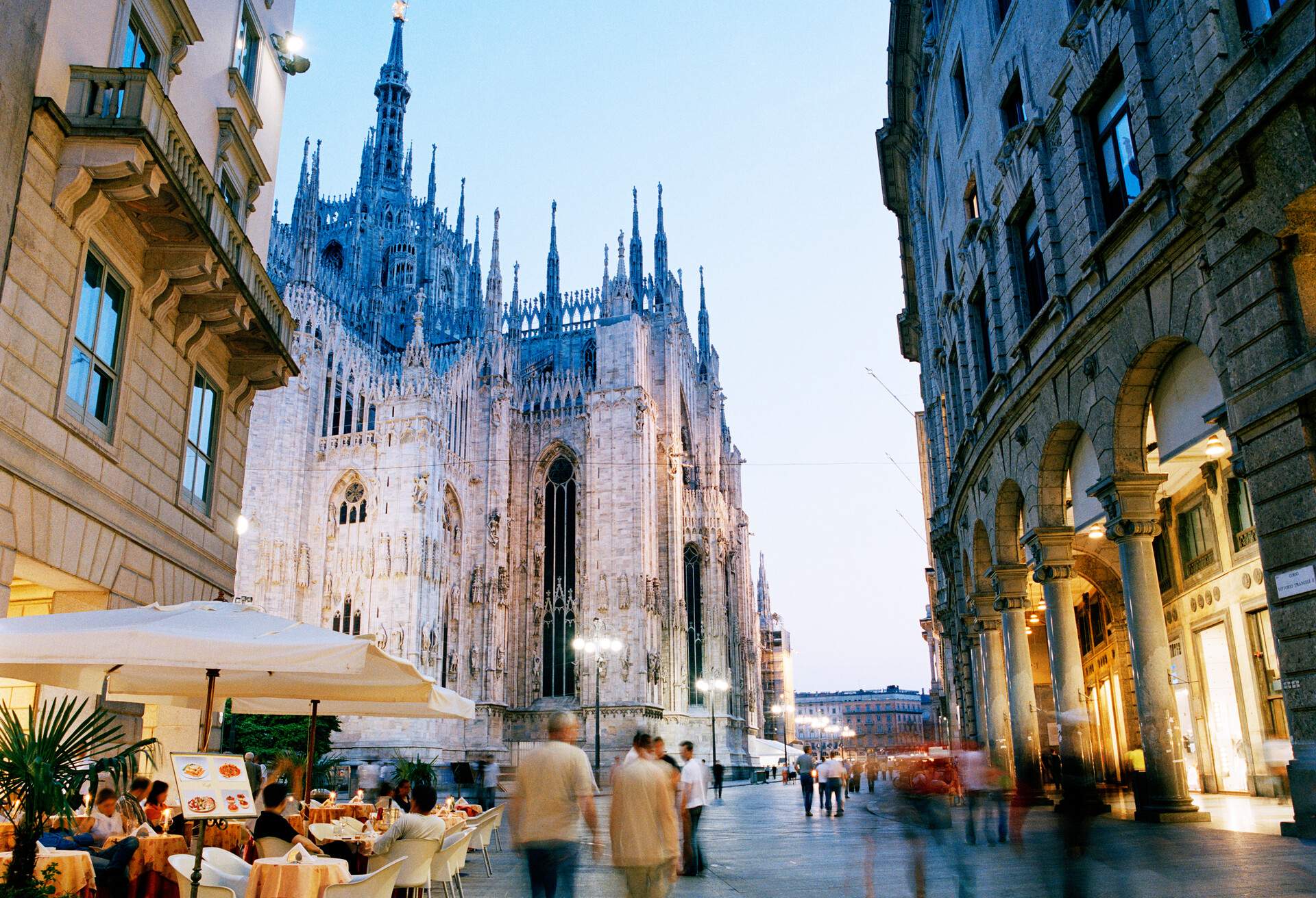 Italy, Lombardy, Milan, Piazza del Duomo, people dining and walking along busy street lined with bars and restaurants in twilight