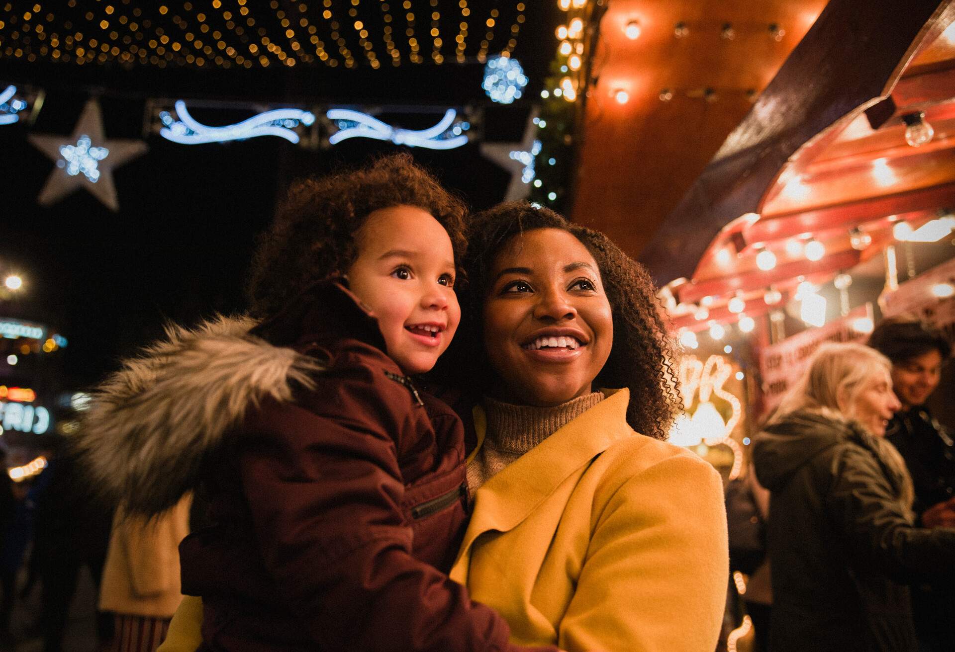 A close-up front-view shot of a woman holding her young daughter, they are both looking at bright​ Christmas lights at the Christmas markets on a cold night, the young girl is in awe.