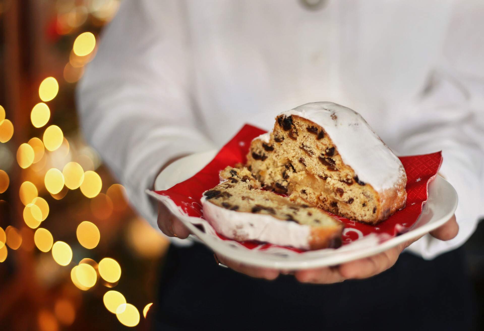 Closeup of a panettone (Christmas sweet) held in the hands of a woman whose body is blurred, dressed in white t-shirt and dark blue pants (trousers) on a background bokeh of Christmas lights.ESPAÑOL: Primer plano de un panetone sostenido en las manos de una mujer cuyo cuerpo se adivina difuminado,  vestida con camisa blanca y pantalón azul oscuro sobre un fondo de bokeh de luces de Navidad.