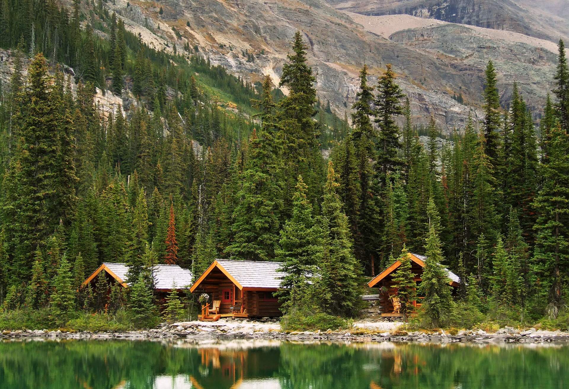Log cabins by the lake with towering trees and rocky mountains.