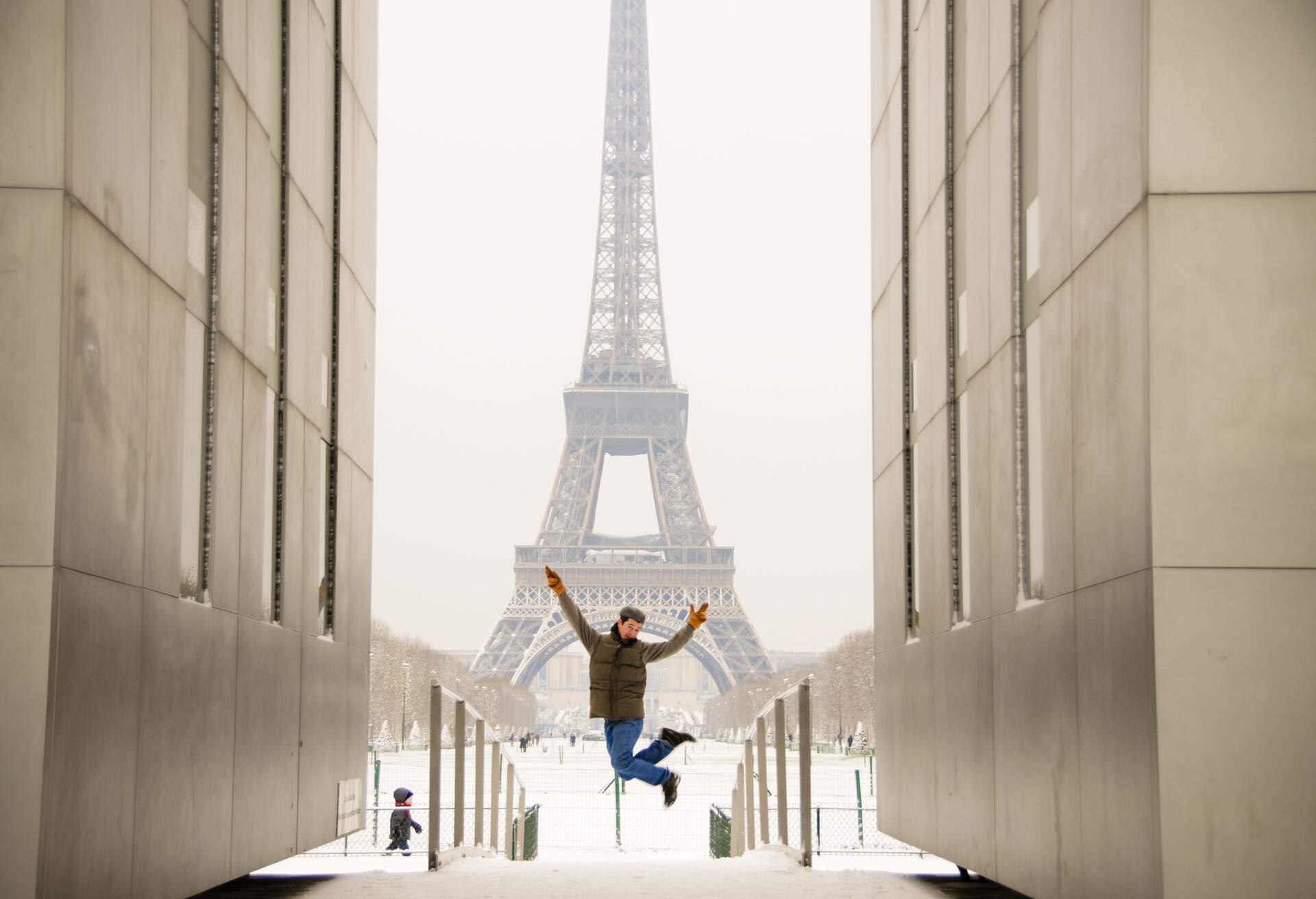 A man in winter jacket jumps between two identical walls against the backdrop of the Eiffel Tower.
