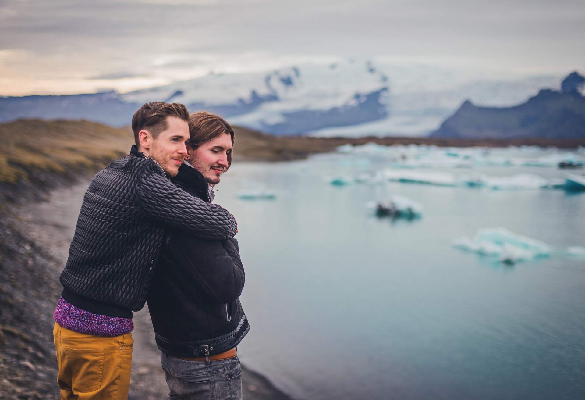 Two young men smiling and holding one another. In the background is Jökulsárlón glacier lagoon/lake in the Vatnajokull region, southern Iceland. Chunks of broken glacier ice float downstream.