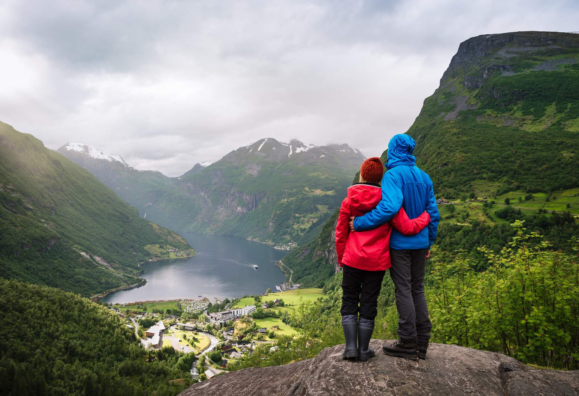 Couple sitting on a rock with looking at the fjord and mountains. Tourist village of Geiranger and the Geirangerfjord, Norway