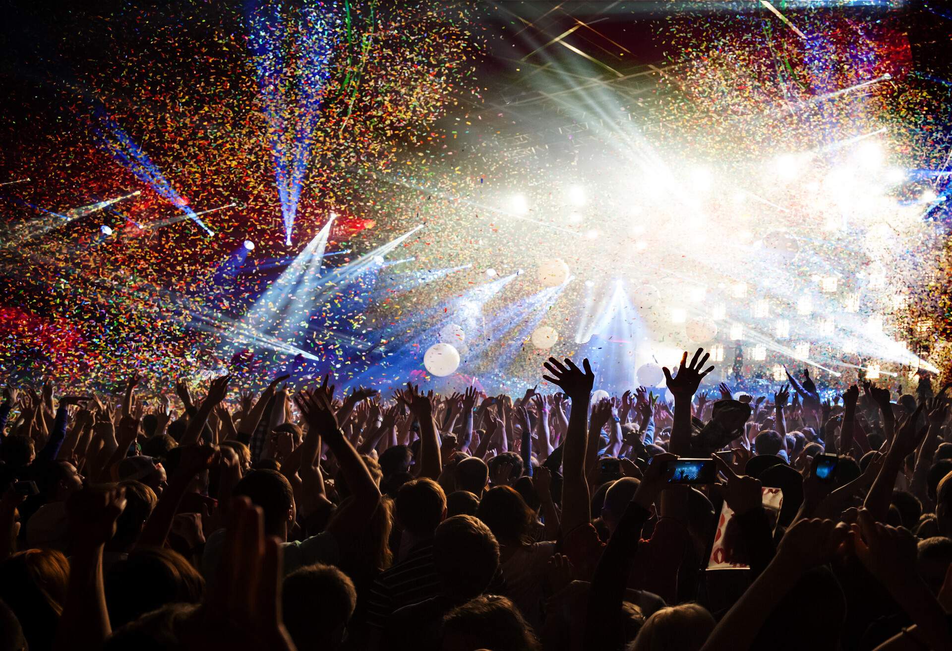 Silhouette of people raising their hands in a concert with glaring stage lights and colourful confetti in the air.