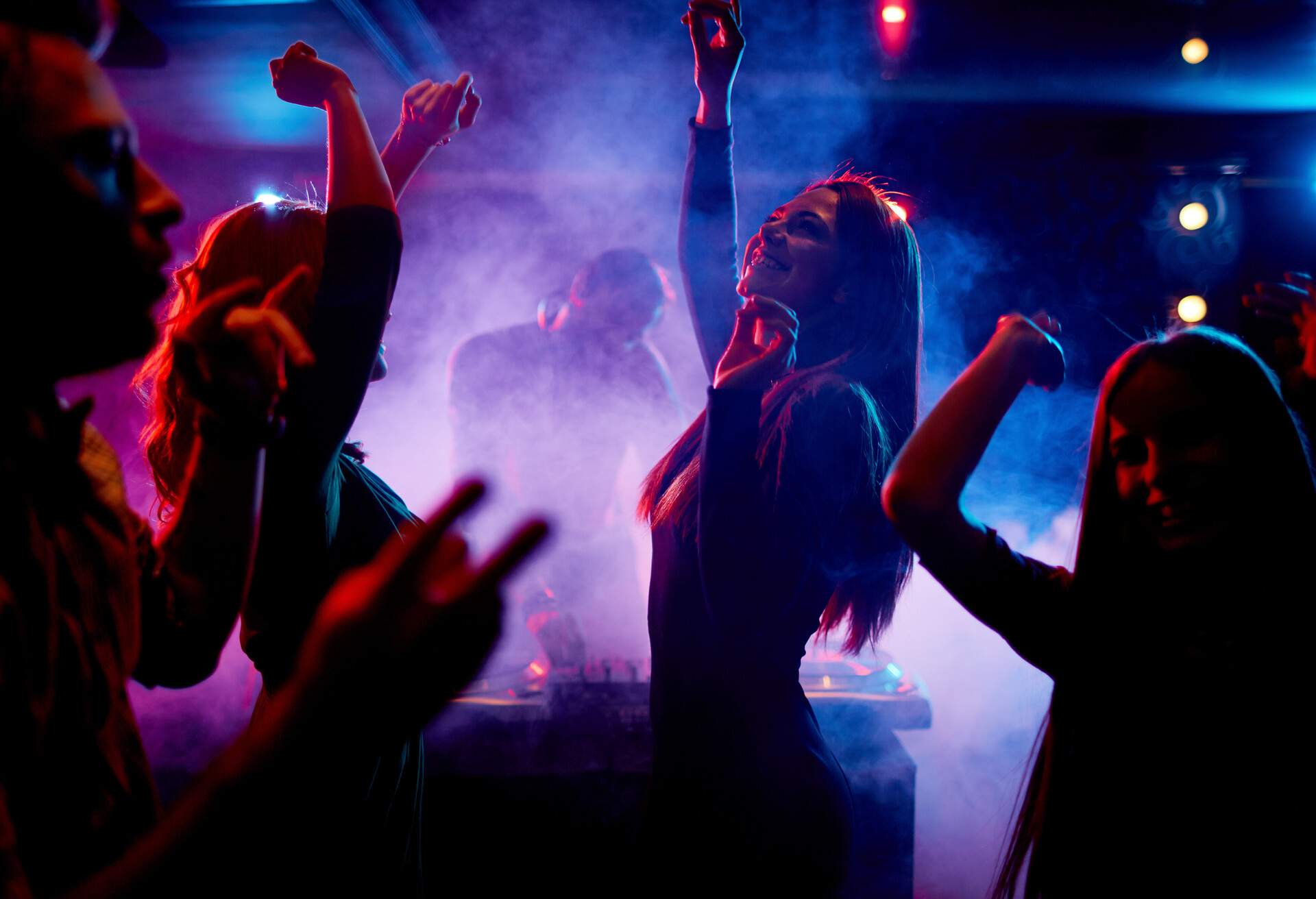 Young people dancing and having a good time in a nightclub while the DJ plays in the background.