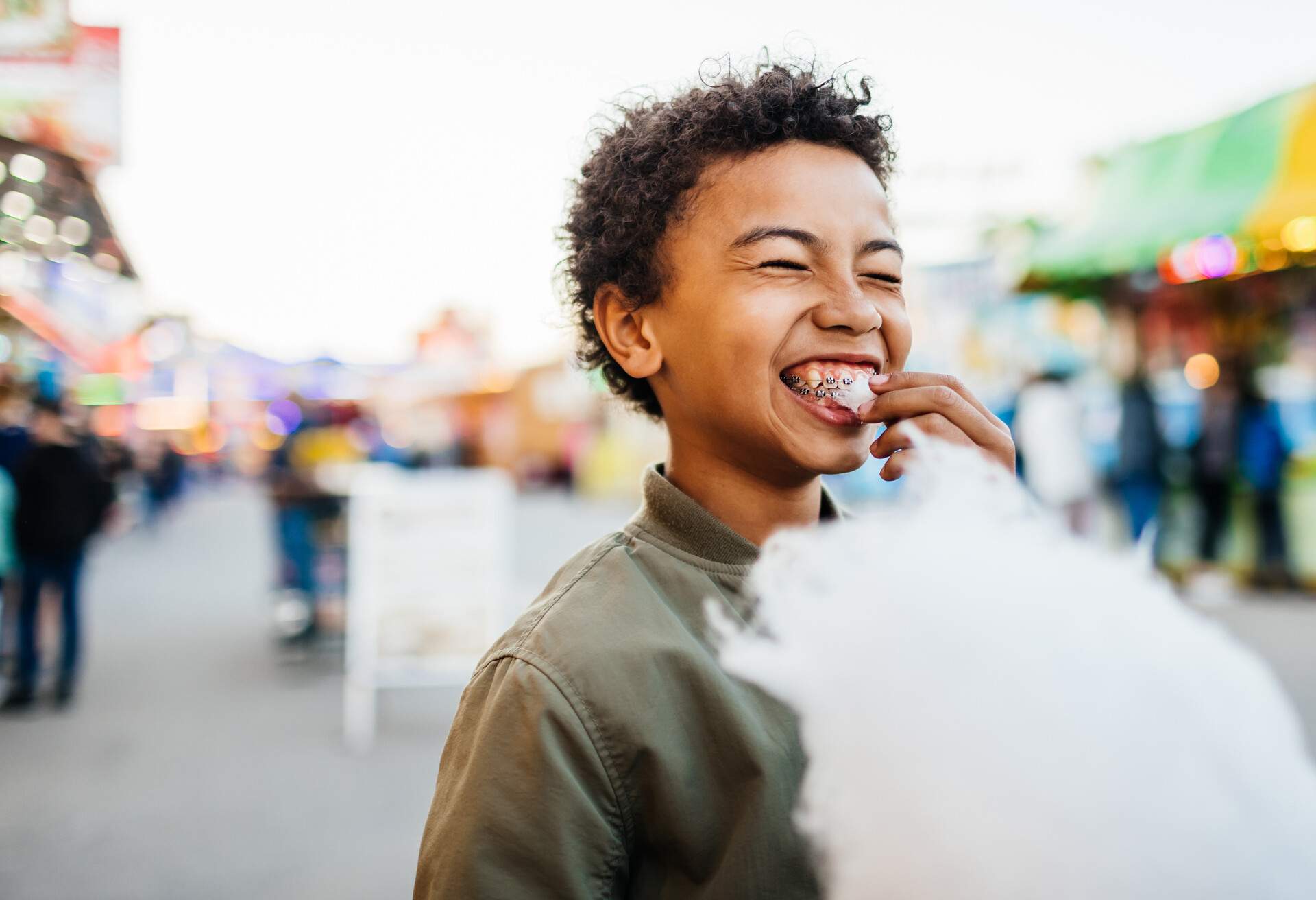 A young boy with braces on his teeth smiling while eating candy floss at the fun fair.