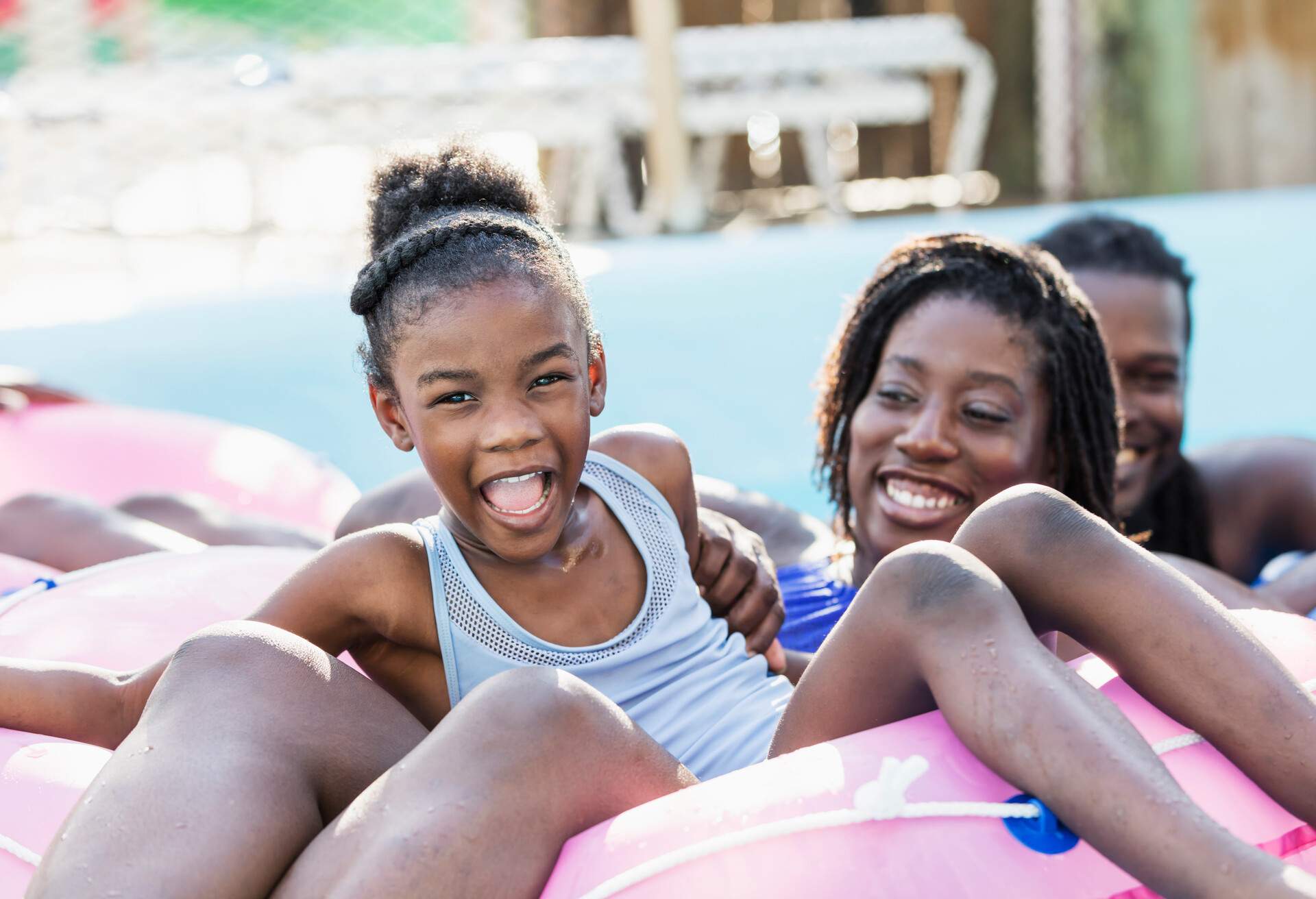 A 6 year old African-American girl having fun at a water park. She and her mother are sitting on an inflatable ring together, floating down a lazy river. She is shouting and laughing, looking at the camera.
