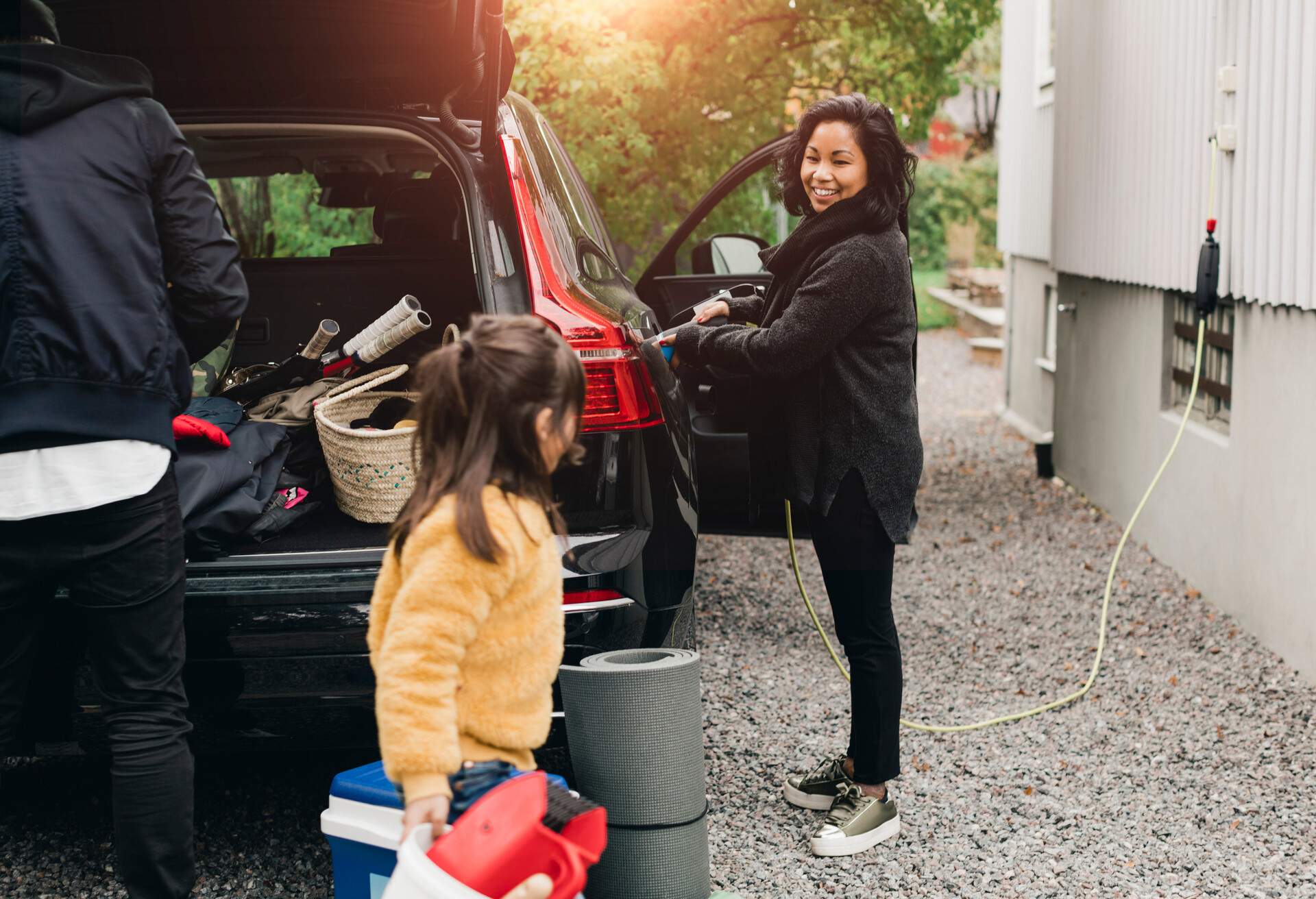Smiling woman charging car while family loading luggage in trunk