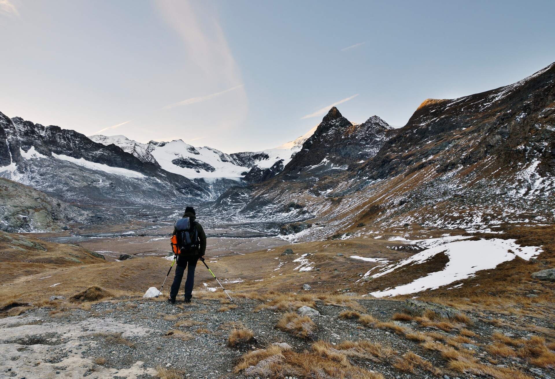 A hiker standing on a dry terrain, looking at a sparsely snow-covered mountain range.