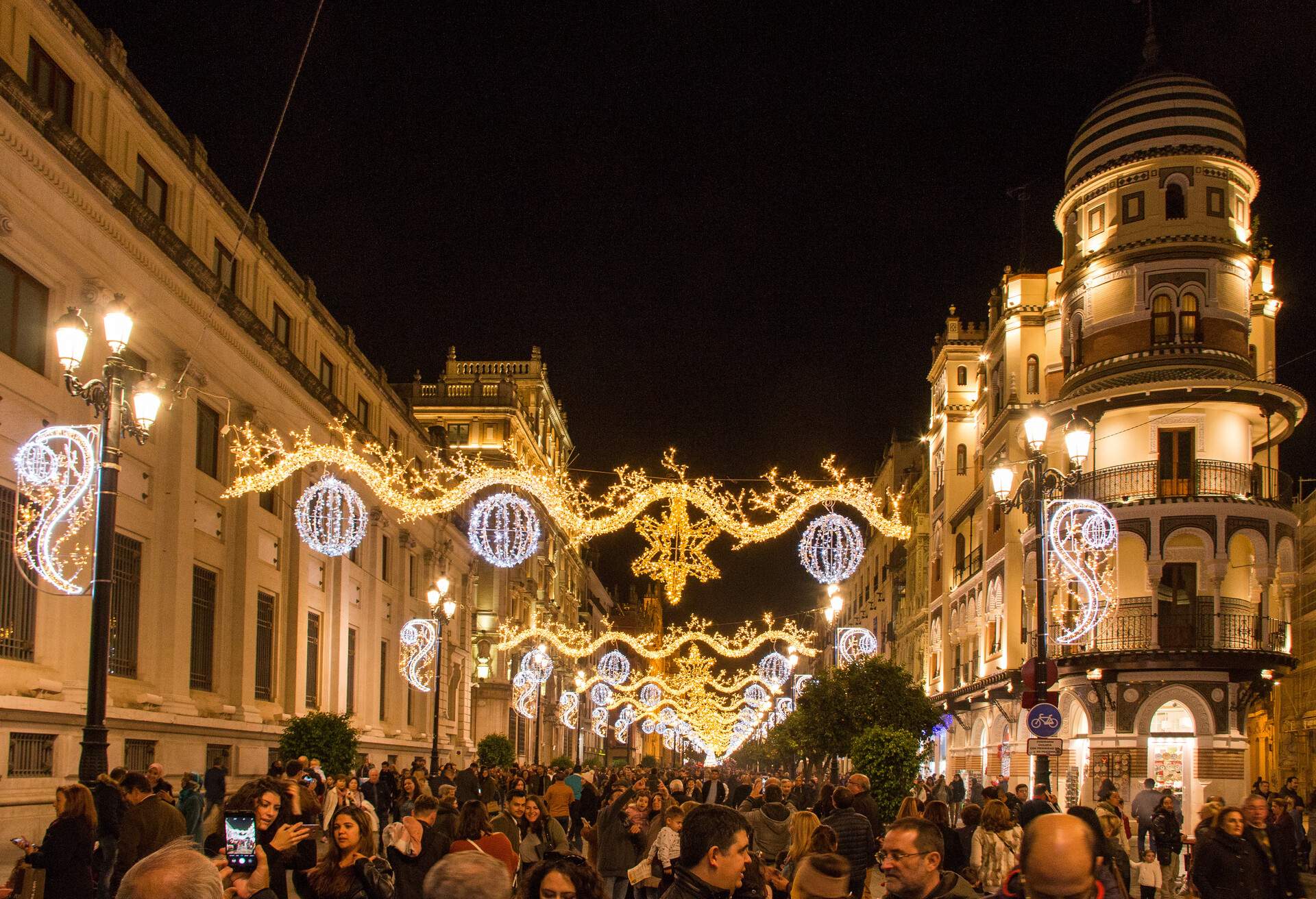 Festive lights hanging over a street lined with elegant structures and packed with people.