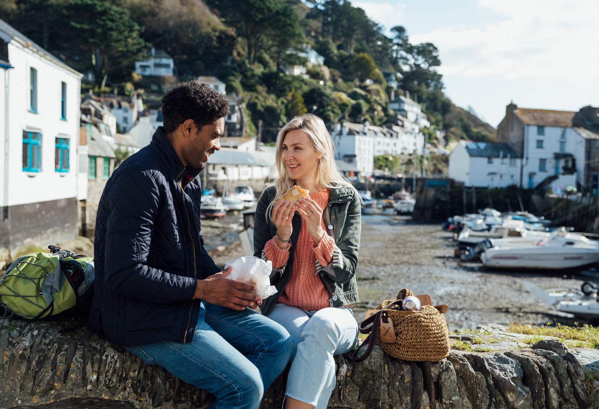 Mid adult couple sitting on a stone wall in Polperro, Cornwall. They are eating Cornish pasties from local businesses while talking with each other.