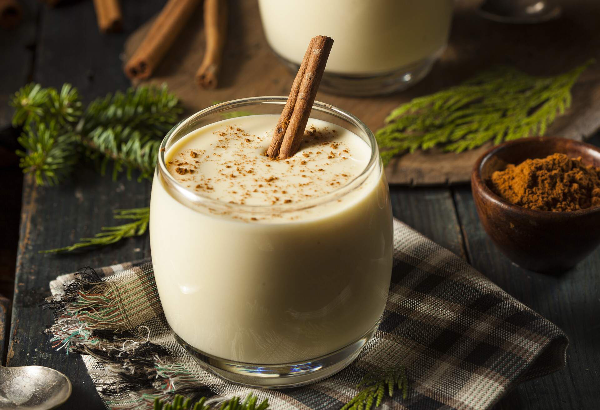 A glass of white foamy drink sprinkled with cinnamon powder and a cinnamon stick, on a wooden table.