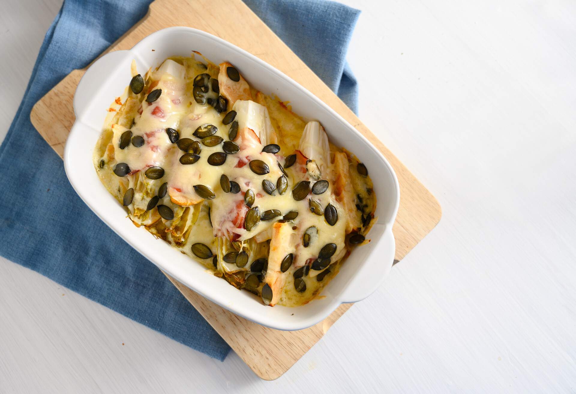 endives or chicory gratin with apple, ham, cheese and pumpkin seeds in a casserole on kitchen board and a blue napkin, white table with copy space, high angle view from above