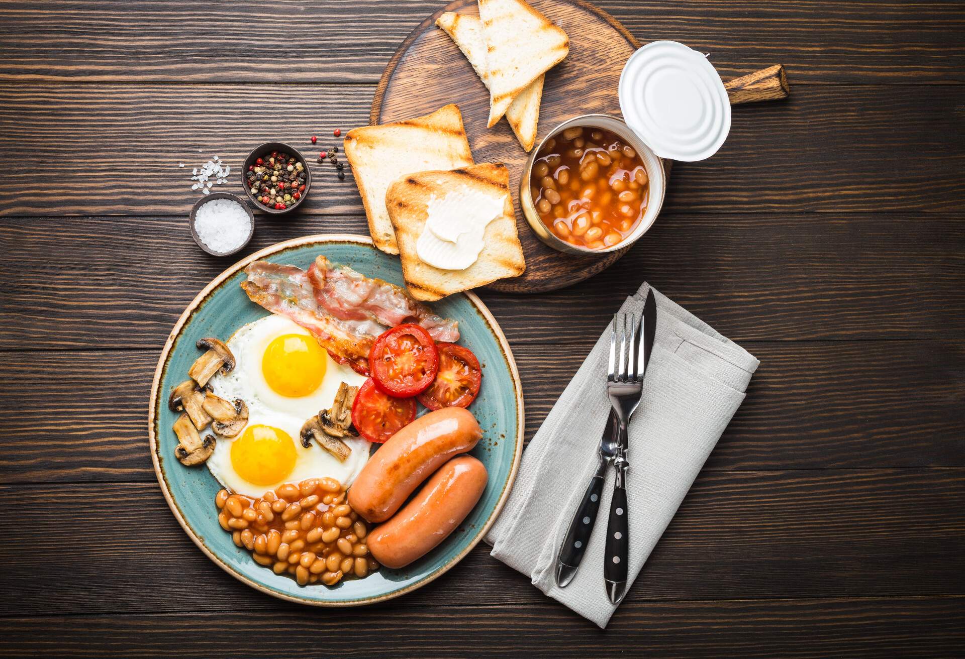Full English breakfast with fried eggs, sausages, bacon, beans, mushrooms, tomatoes on a plate, bread toasts with butter. Traditional British meal, top view, rustic wooden background