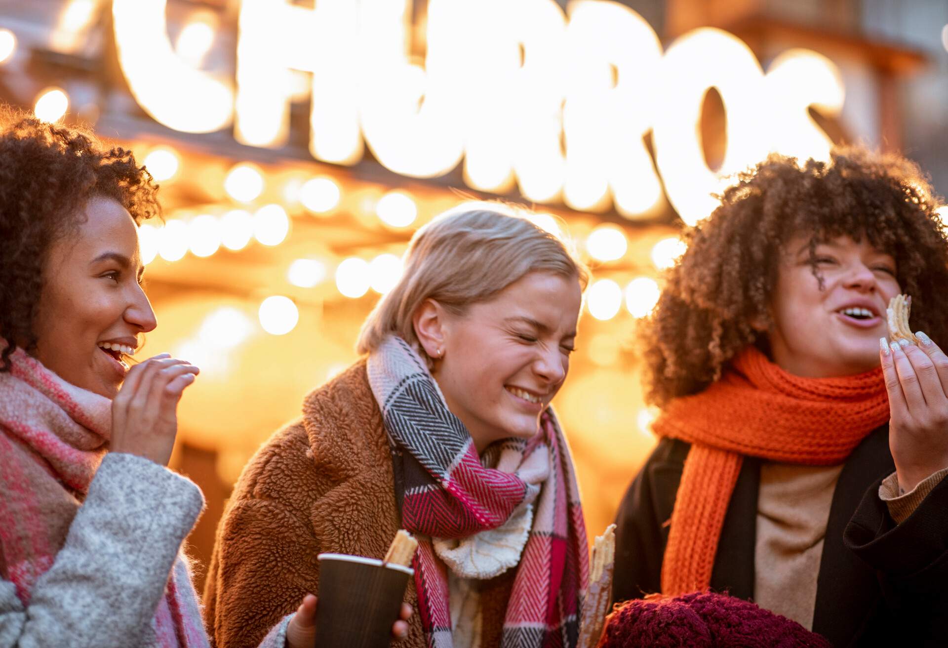 Three women in winter clothes laughing while eating churros.
