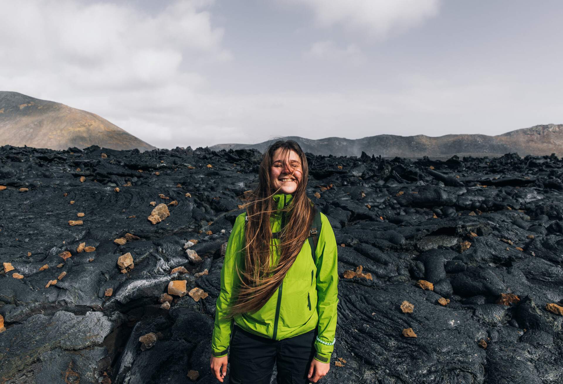Young smiling woman explorer with long hair onin green jacket exploring the dramatic landscape of the new erupting volcano with the flame melting lava