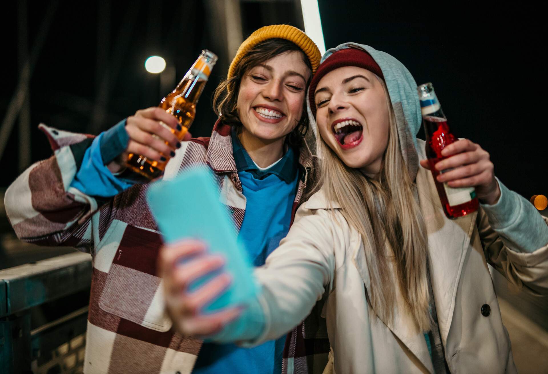 Spontaneous photo of two beautiful, cute women, a brunet and blonde, drinking alcohol, cheering and having fun while taking a selfie, on the street at night.