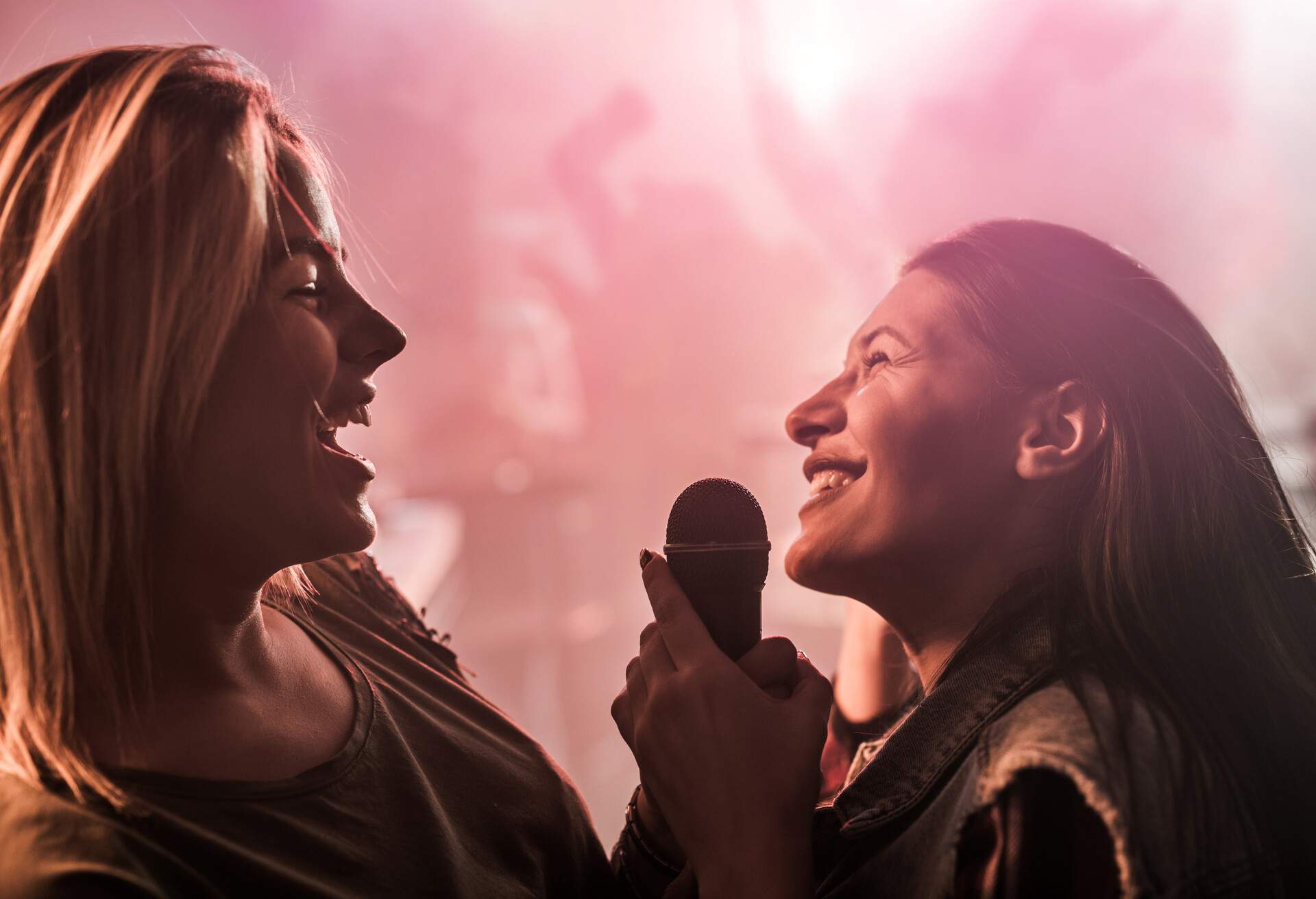 Happy female friends holding microphone and singing at karaoke party in nightclub. There are people dancing in the background.