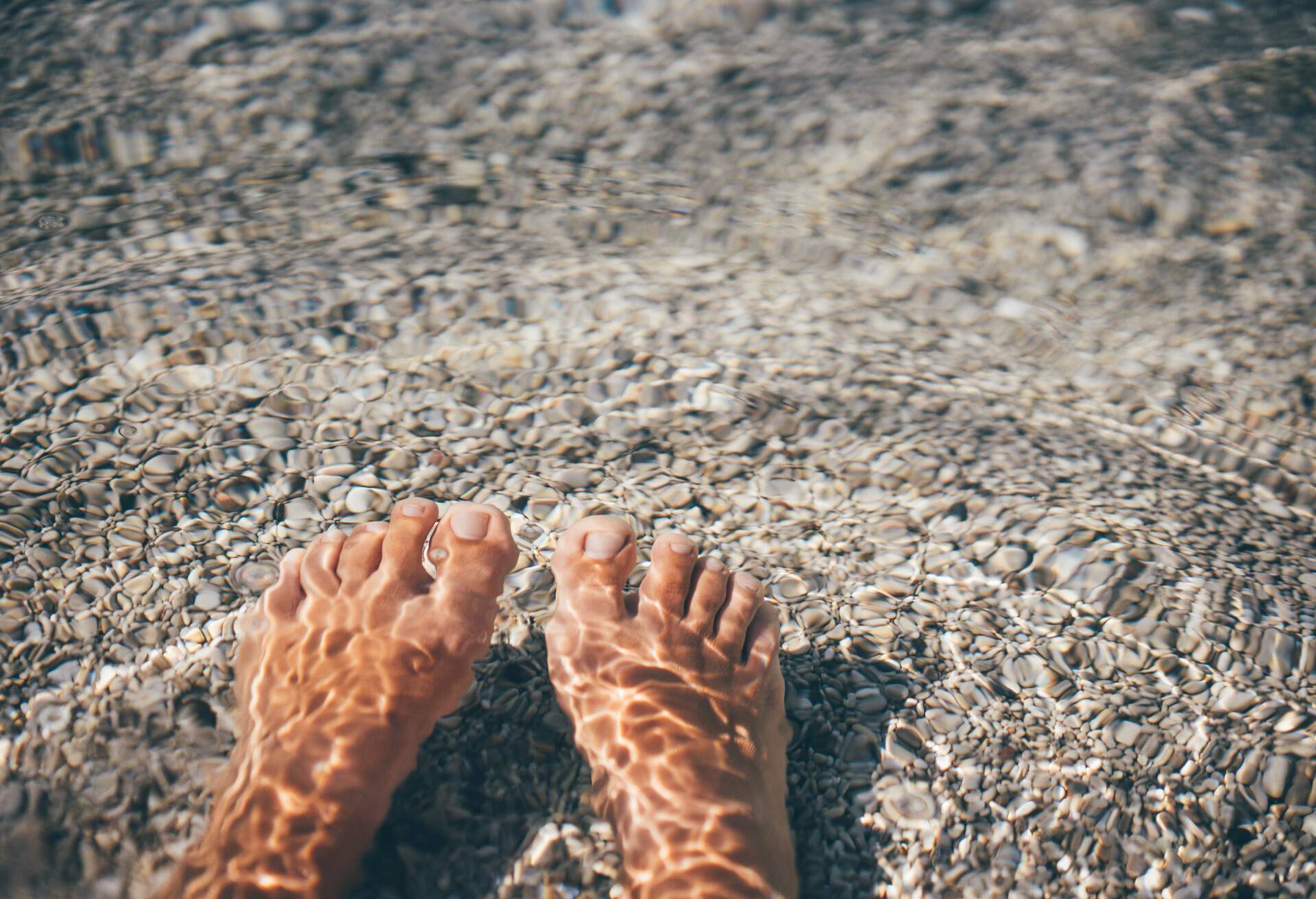 A woman's feet submerged in a crystal-clear sea with a pebbly bottom.