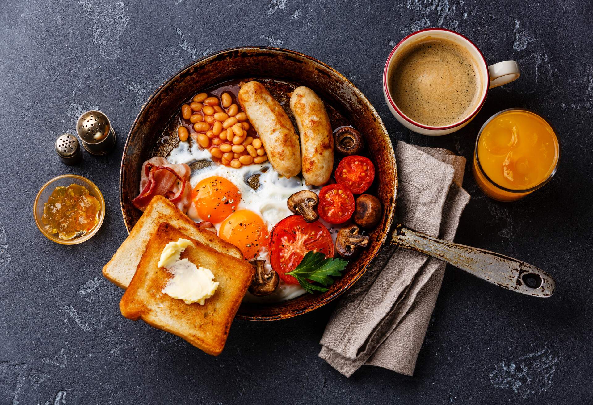 English breakfast in pan with fried eggs, sausages, bacon, beans, toasts and coffee on dark stone background; Shutterstock ID 558715117