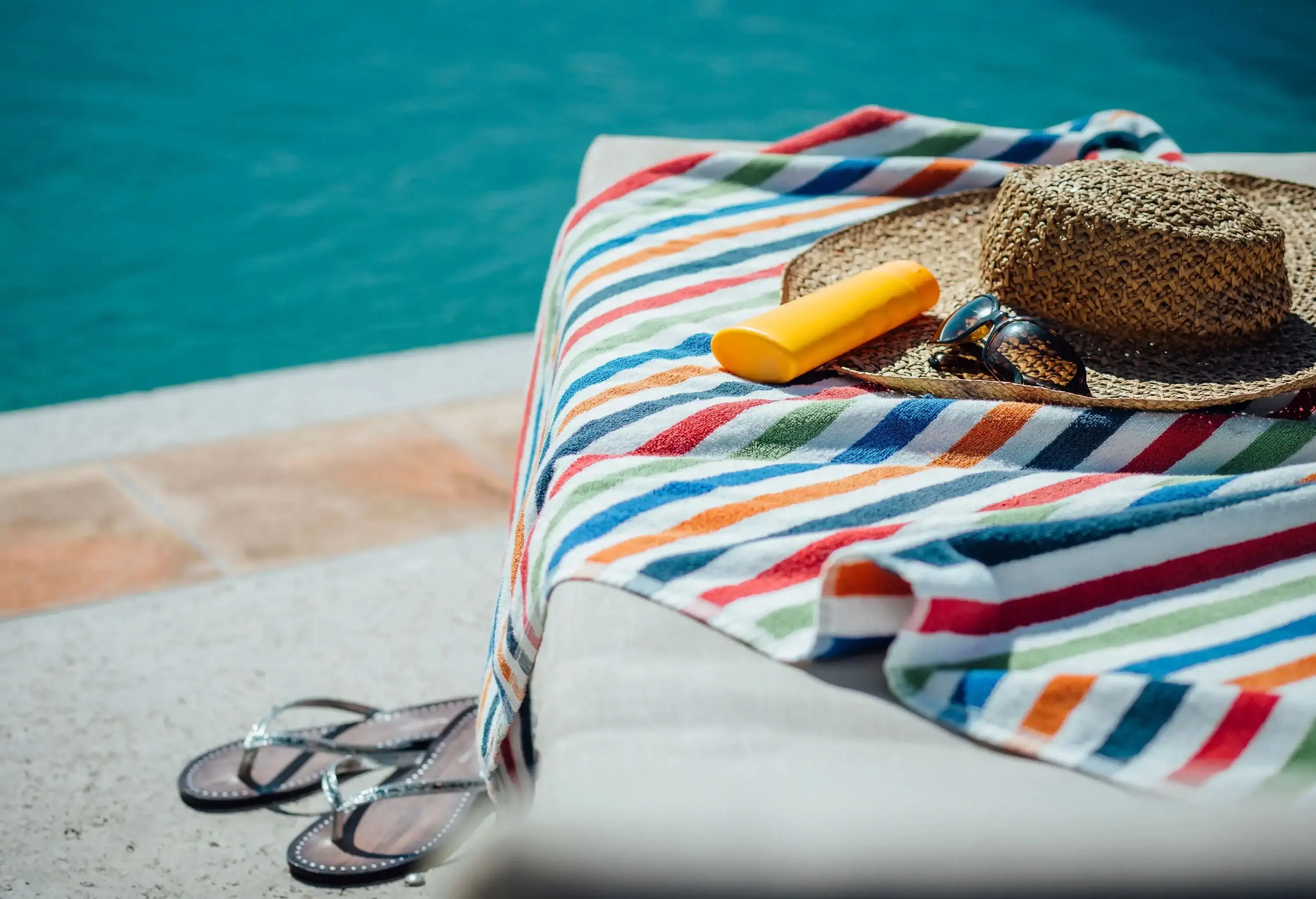 A colourful stripe towel, summer hat, sunglasses, yellow case, and slippers on a sunlounger next to the pool.