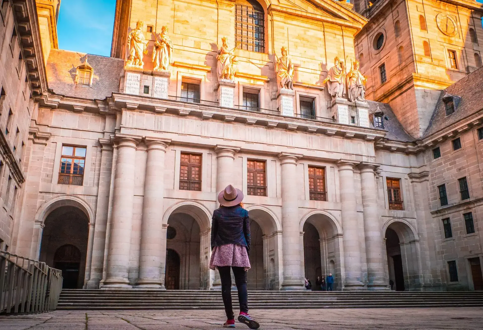 A woman stands in front of a basilica with massive white columns and arched entrances, looking up at the sun-lit human sculptures.