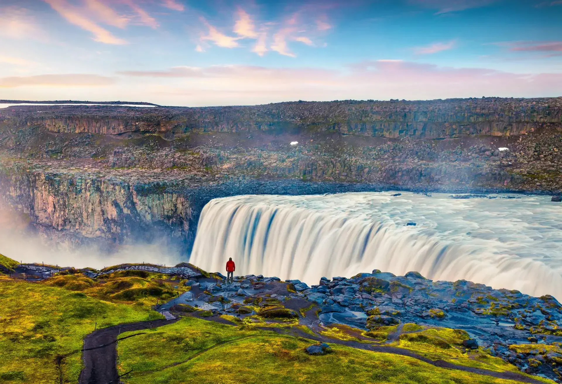 Tourist admiring view of falling water of the most powerful waterfall in Europe - Dettifoss. Colorful summer sunrise in Jokulsargljufur National Park, Iceland. Artistic style post processed photo.