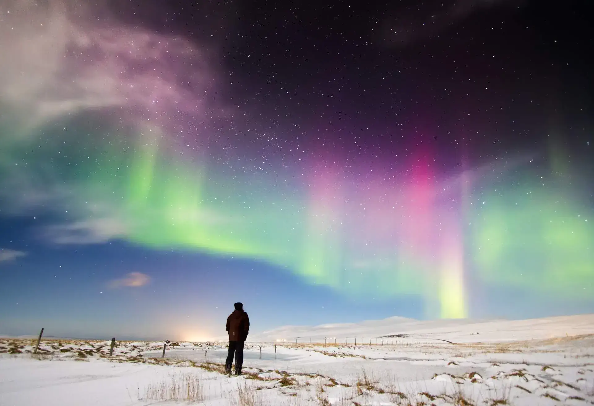 A man standing on a snowfield gazing at the northern lights in a night sky filled with stars.