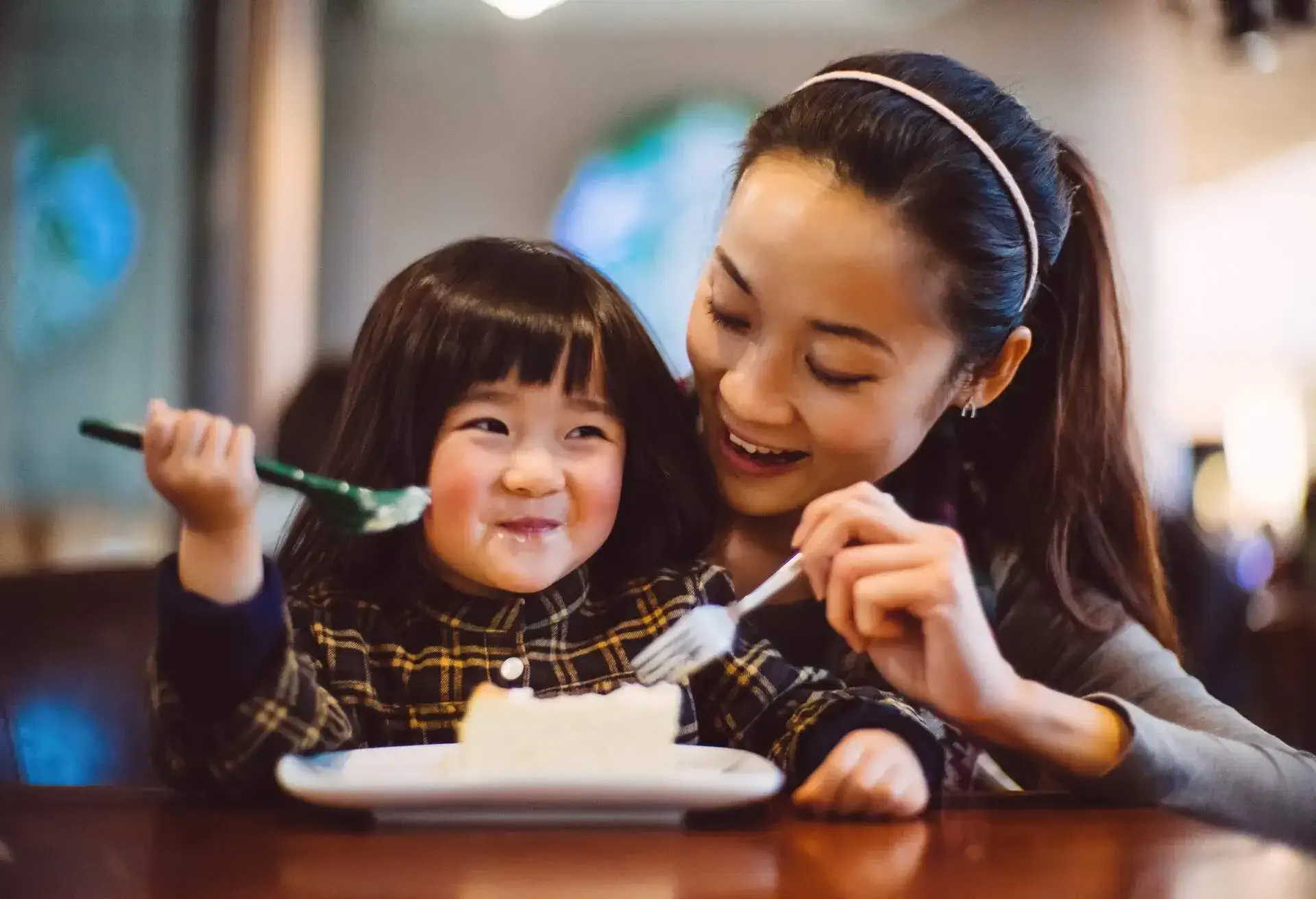 Pretty young mom & lovely toddler girl having cheese cake while talking to each other joyfully in the cafe