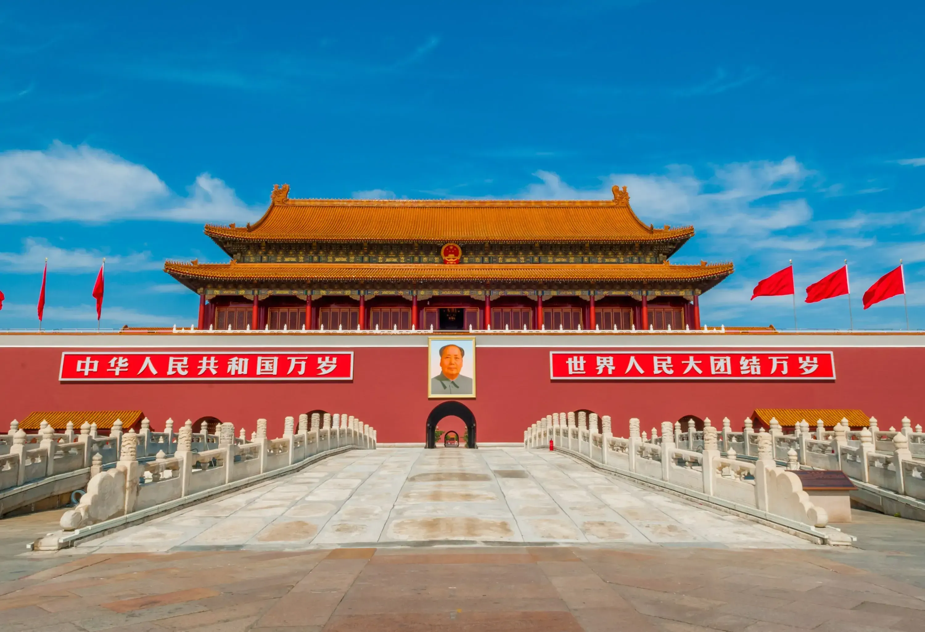 A stone bridge leads across a wide pathway to a majestic traditional building with golden-tiled roof, red walls, and a large central portrait of a person on a bright day.