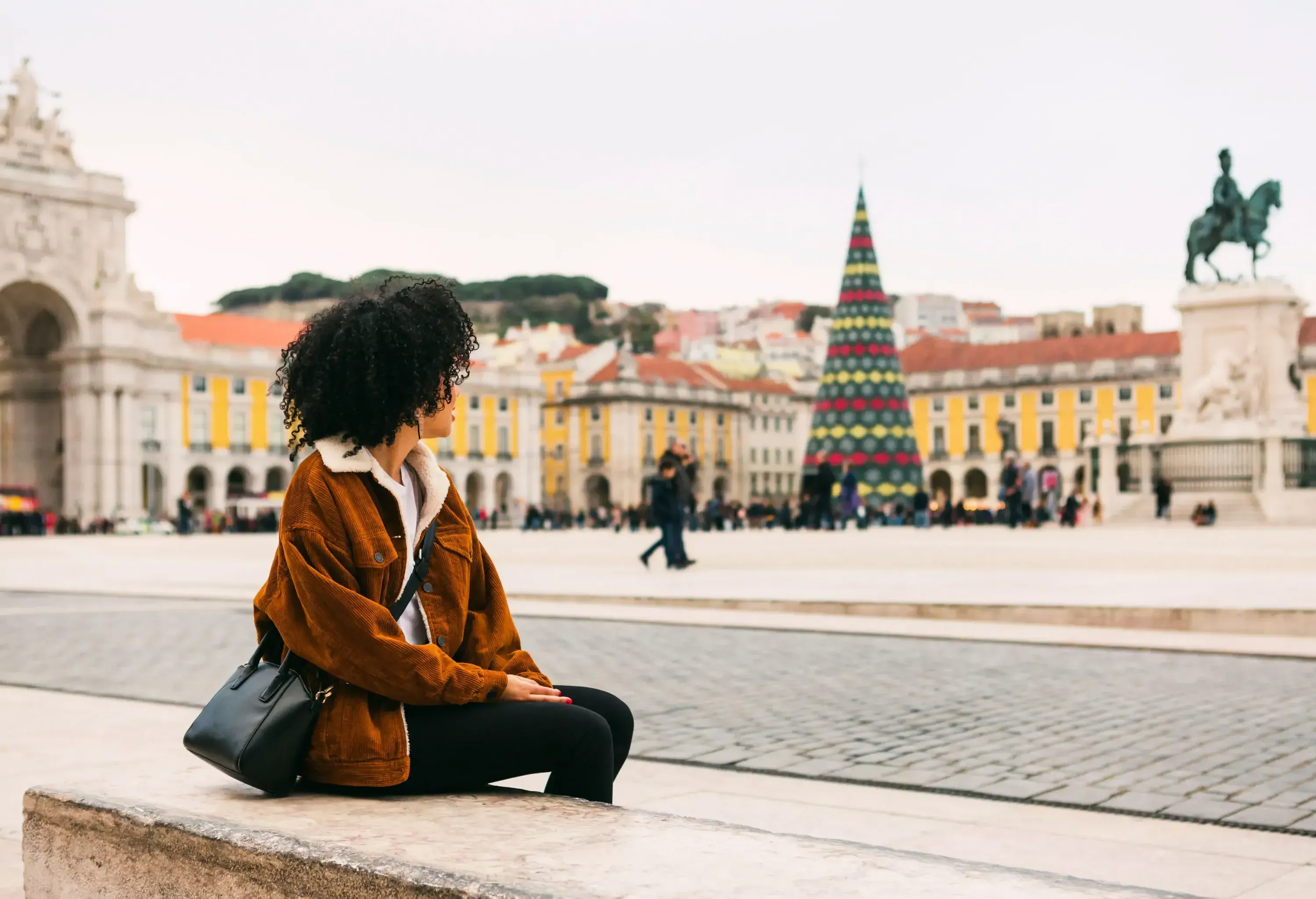 A person is sitting by a walkway, looking at a Christmas tree on a town square.