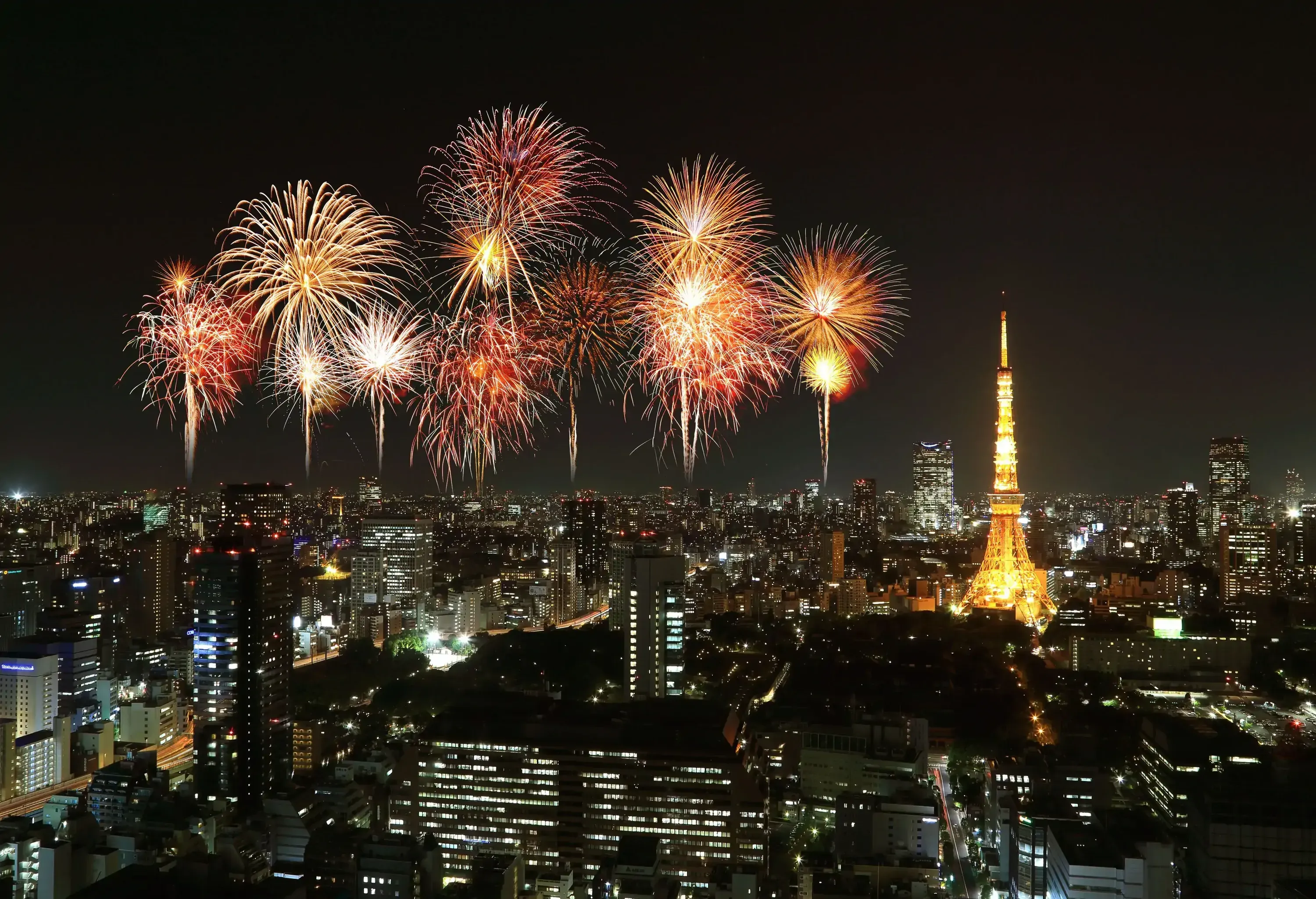 Vibrant, colourful fireworks explode dazzling in the night sky behind an illuminated tower in the middle of a cityscape.