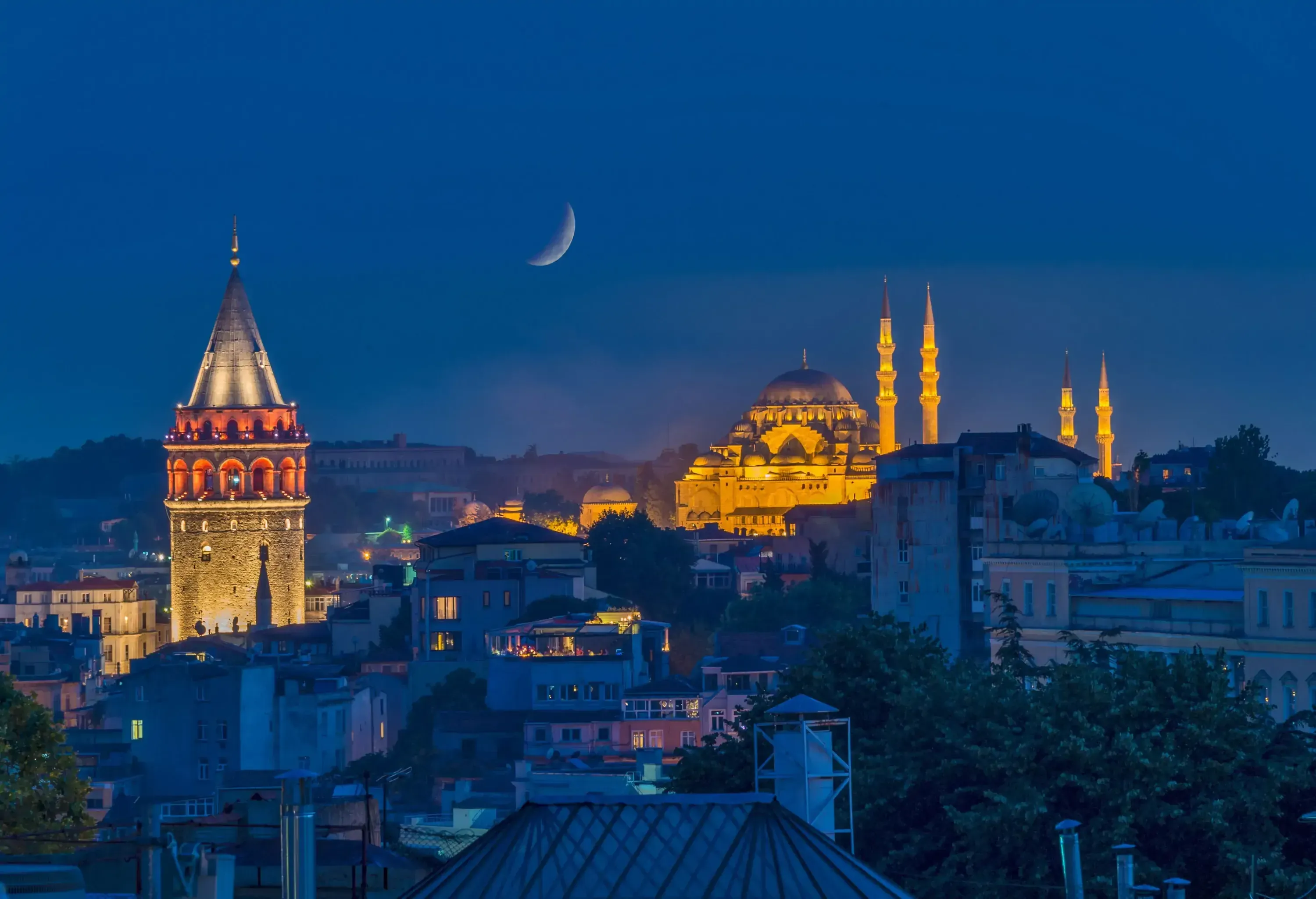 A night-time view of the illuminated Galata Tower and Süleymaniye Mosque rising above a cityscape under a crescent moon.
