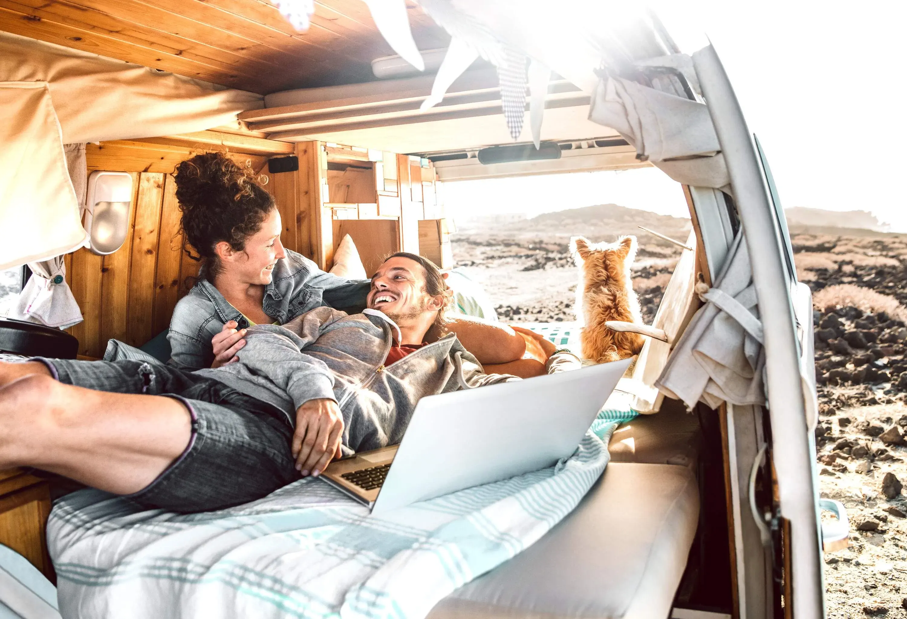 Couple grinning at each other as they lie on a bed inside the van with a dog gazing out the window.