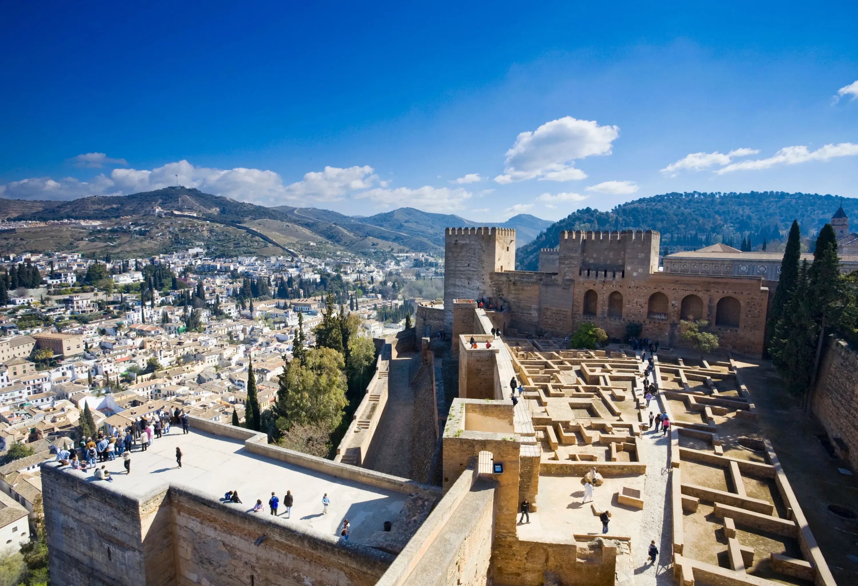 A wide view of an ancient stone fortress and its observers overlooking a city nestled among hills under a blue sky.