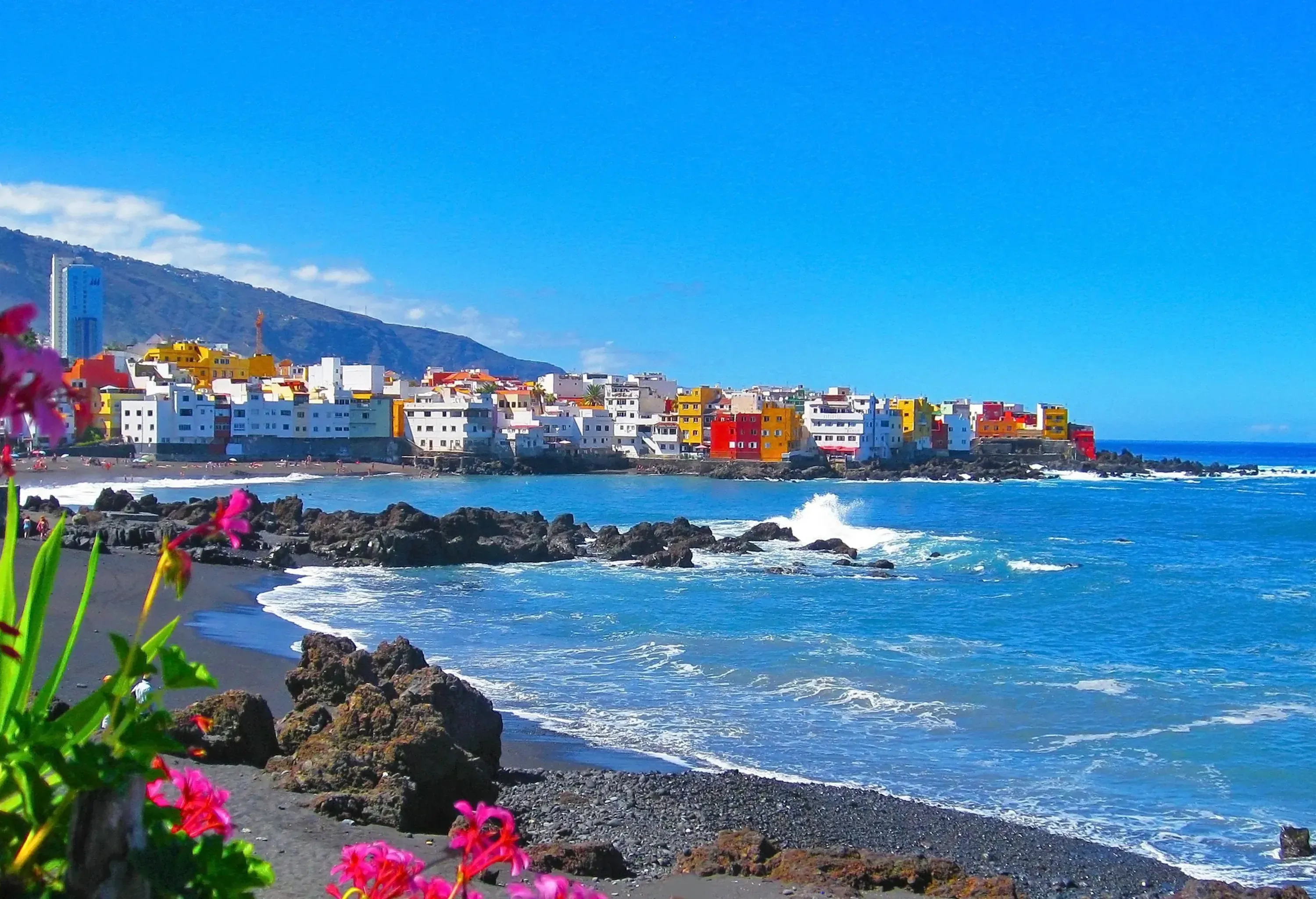 Waves crashing against the rocks on the beach, with colourful buildings in the backdrop.