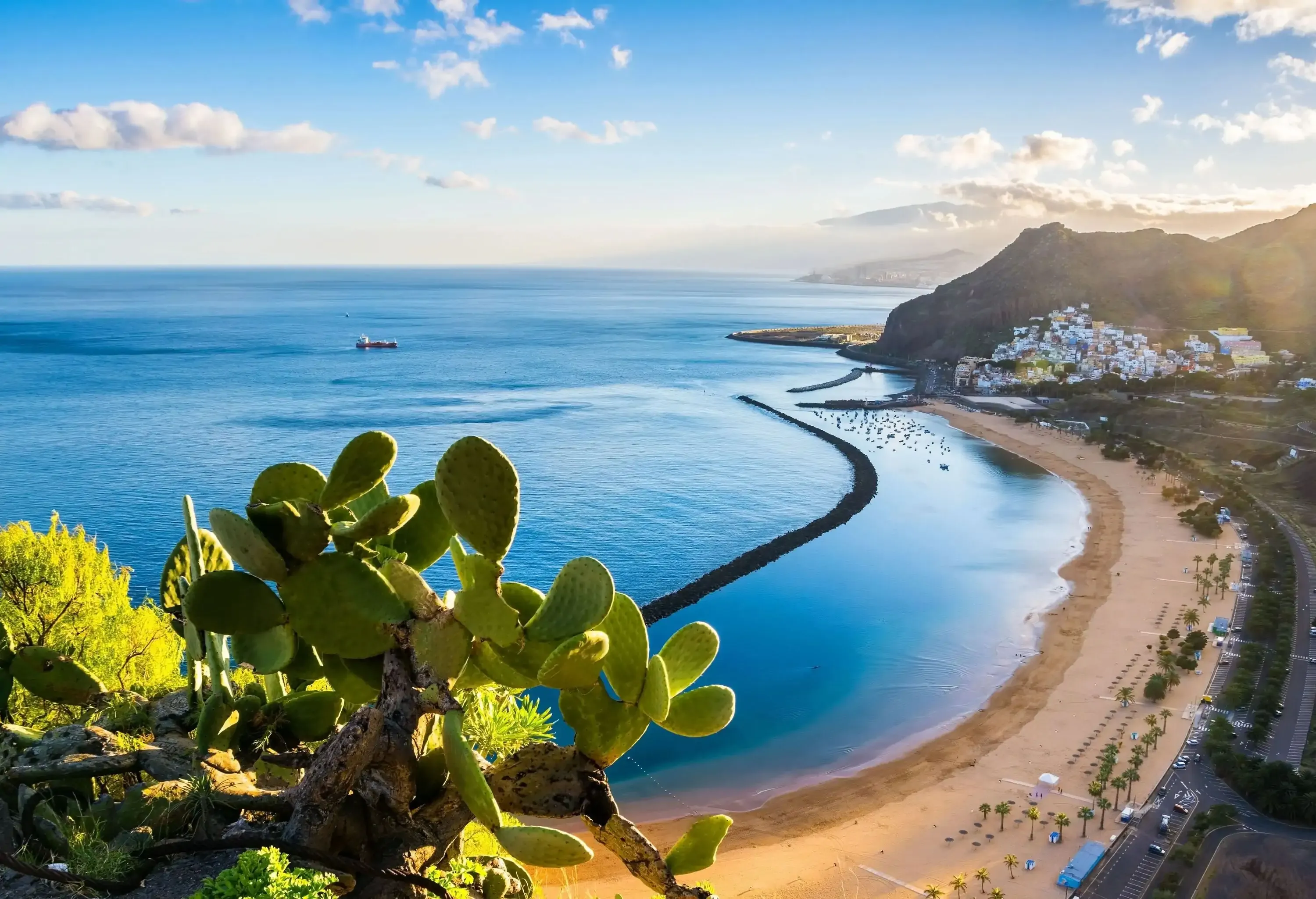 A winding road with a beautiful, breakwater-protected beach and a hilltop settlement in the distance.